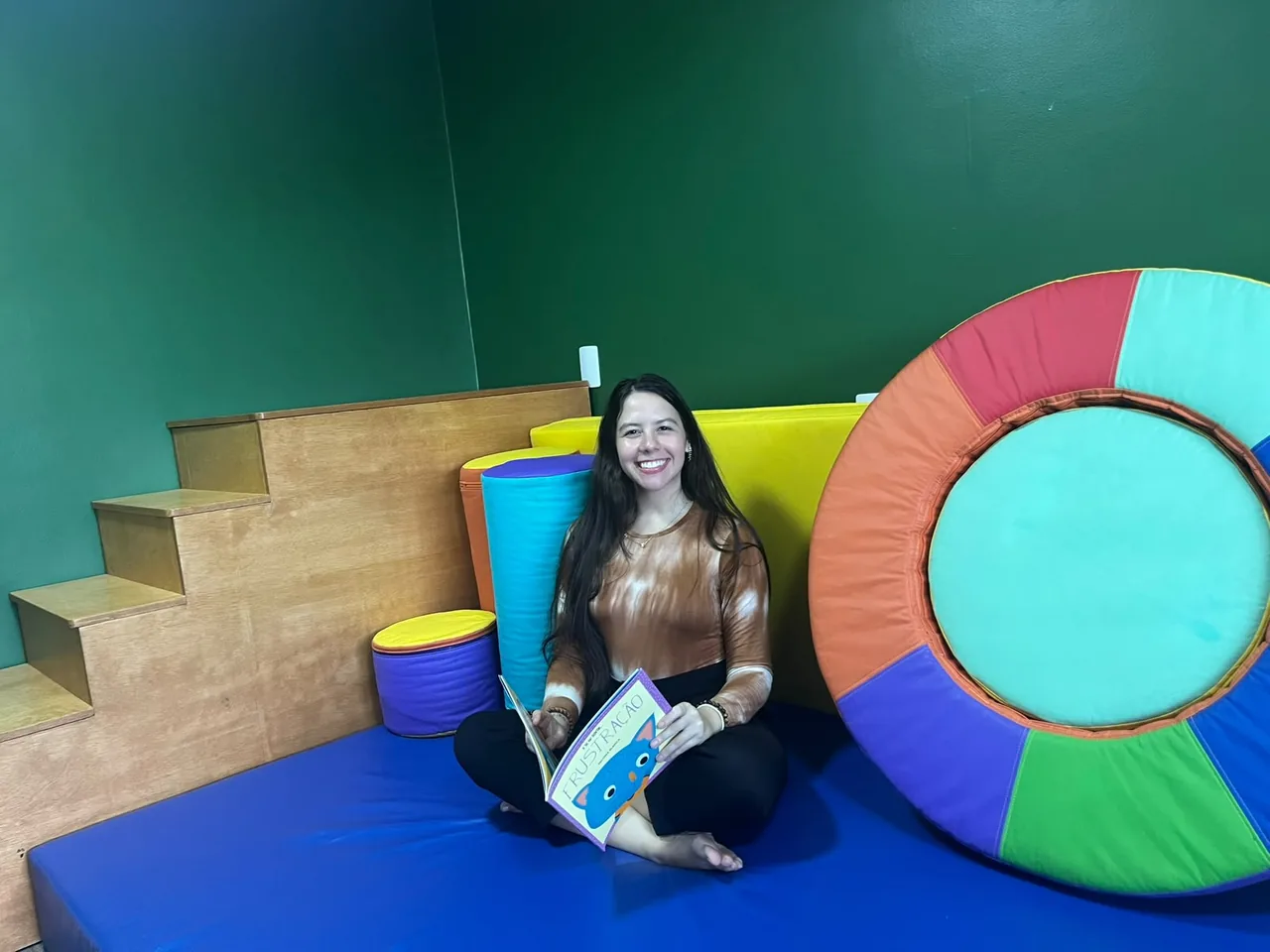Maria Eduarda Mendonça lendo um livro infantil sobre frustração no espaço lúdico do consultório, cercada por almofadas e estruturas coloridas